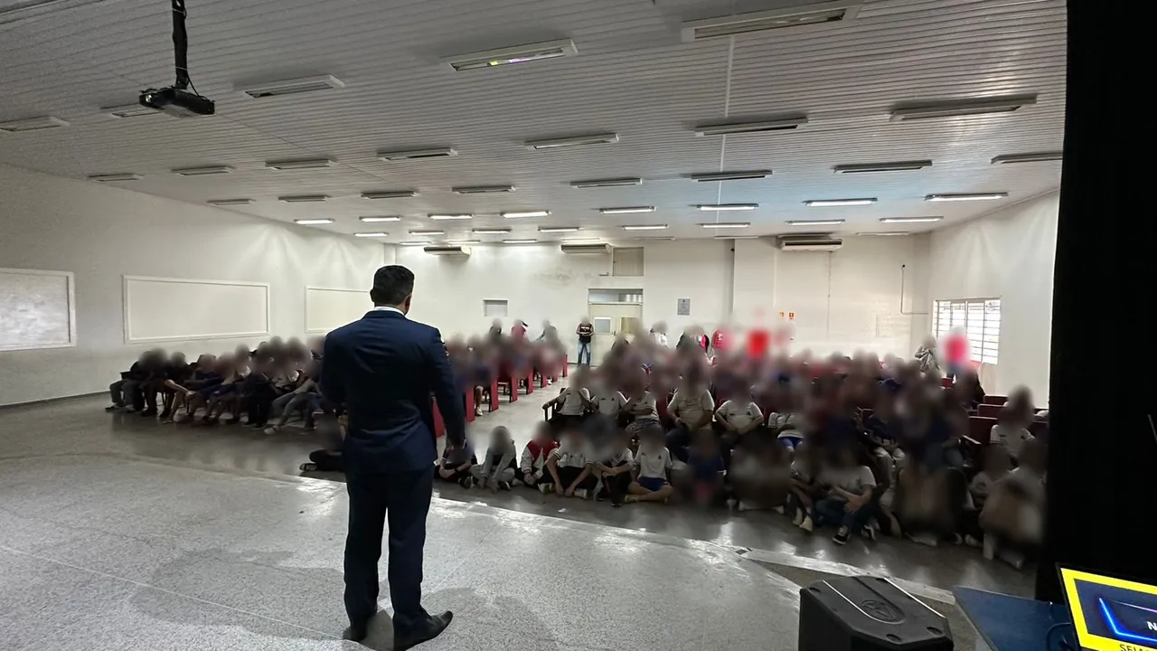 Anderson Elias, de costas e de terno azul-marinho, fala do palco para um auditório lotado de estudantes. Os rostos da plateia estão desfocados para preservar a identidade dos alunos.