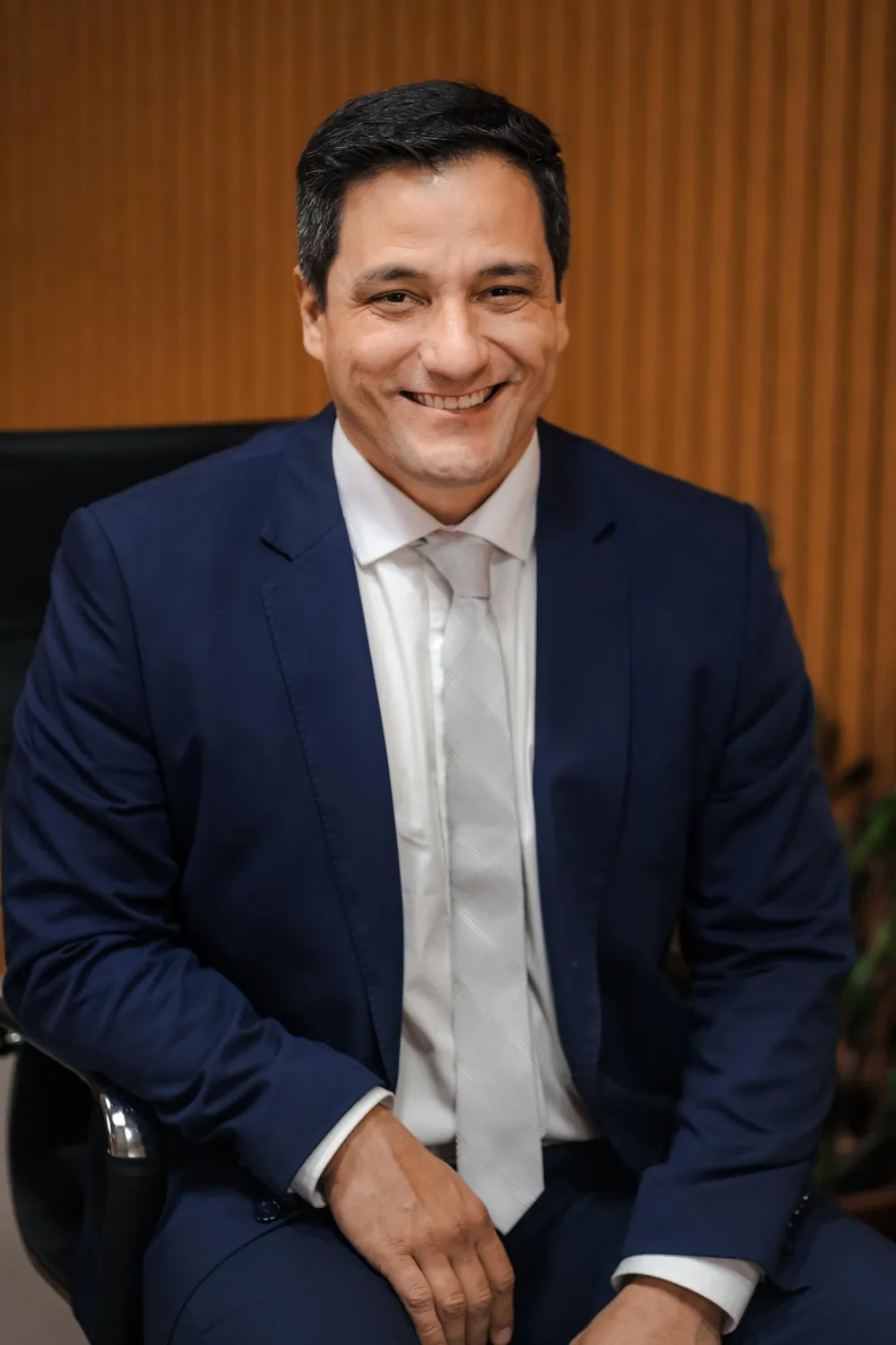 Anderson Elias de terno azul-marinho, sentado e sorrindo, diante de uma parede de ripas de madeira.