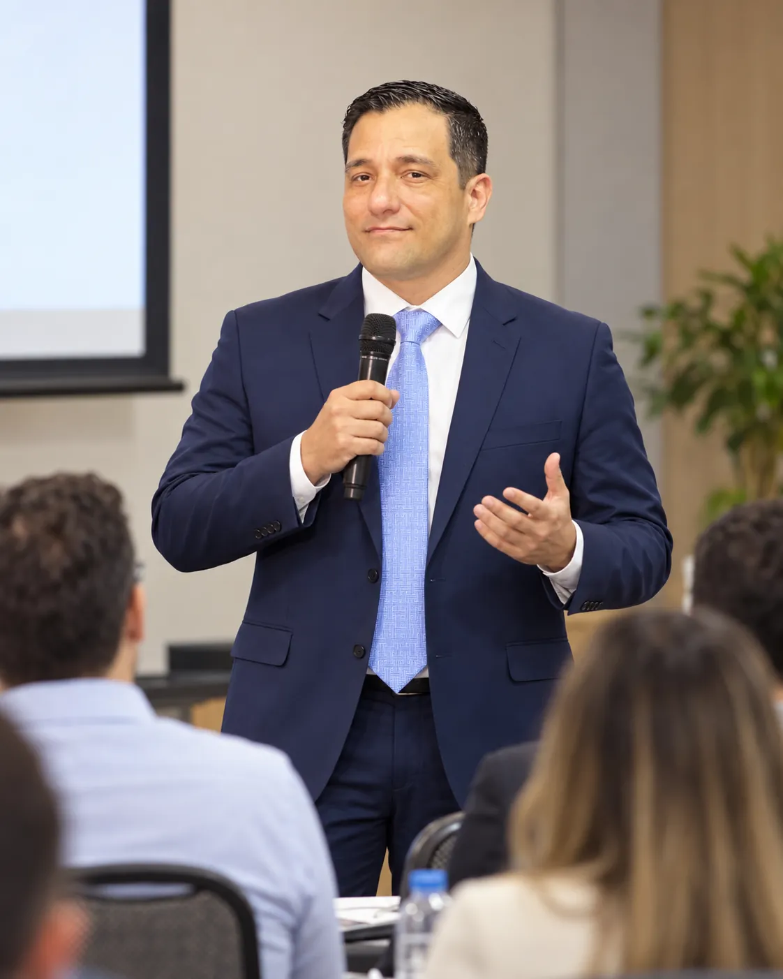 Anderson Elias, de terno azul-marinho e gravata azul-clara, fala ao microfone para uma plateia sentada em uma sala de reuniões.